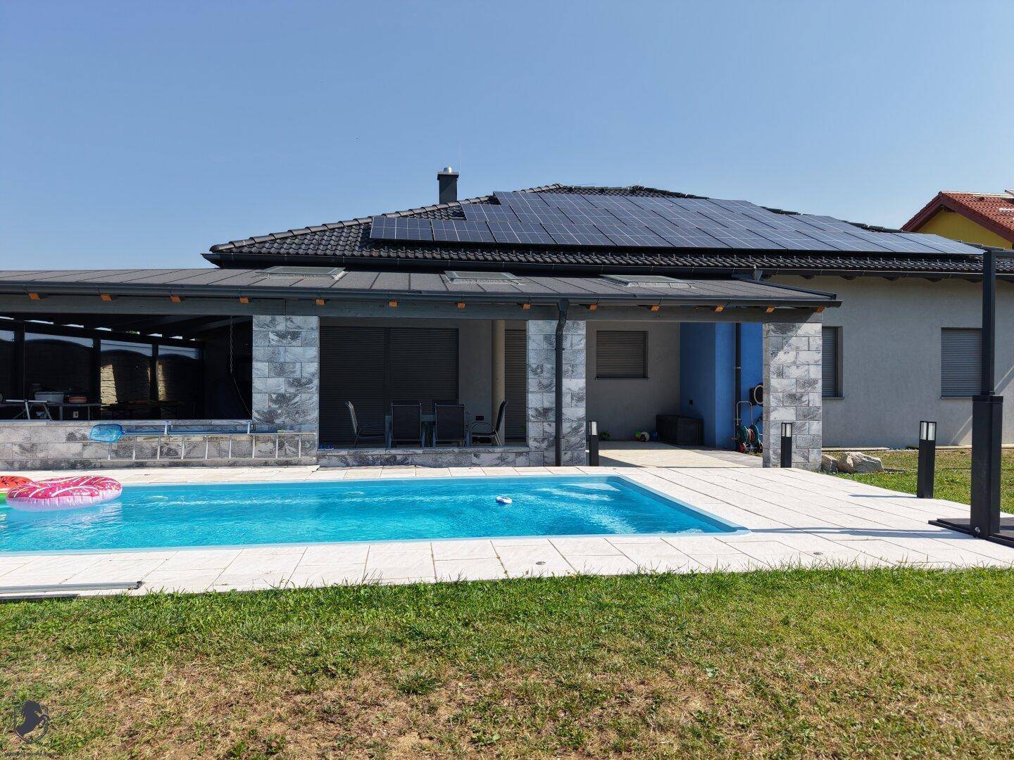Außenbereich mit großem Pool, überdachter Terrasse und dem Haus mit Solaranlagen im Hintergrund.