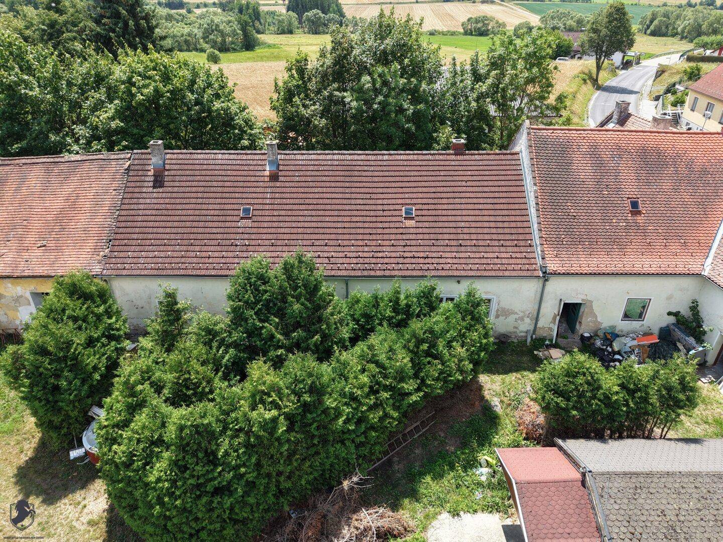 Langes Gebäude mit rotem Ziegeldach, umgeben von dichten Hecken und Bäumen im ländlichen Umfeld.