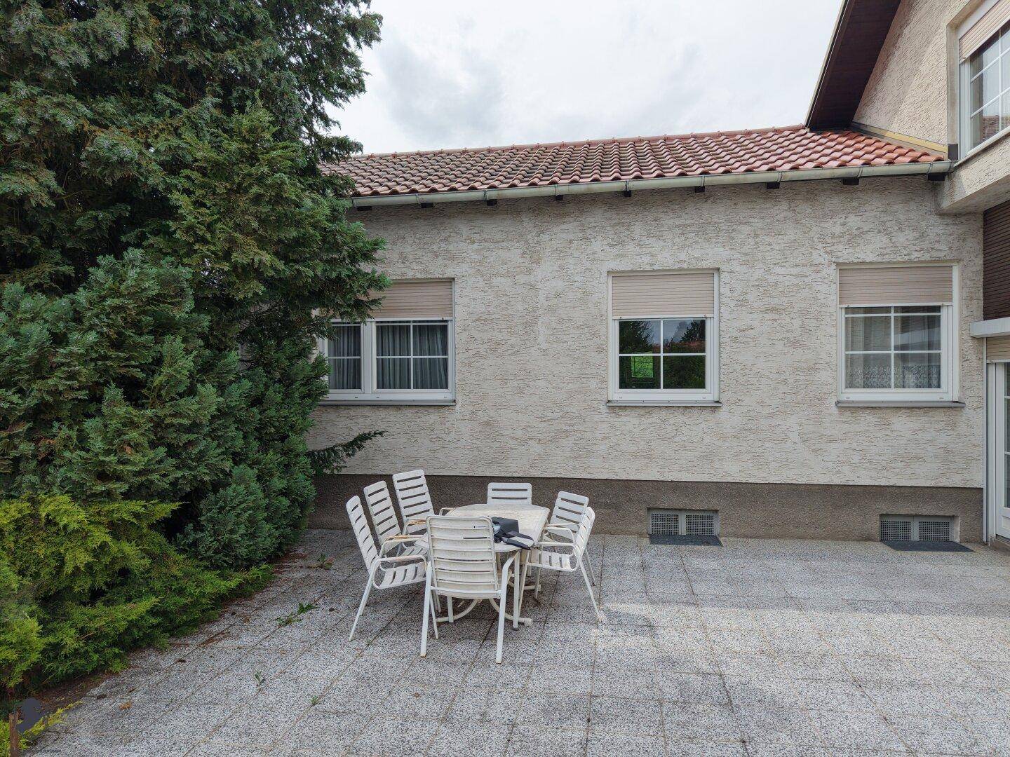 Gepflasterte Terrasse mit Gartenmöbeln neben dem Haus, umgeben von Bäumen.