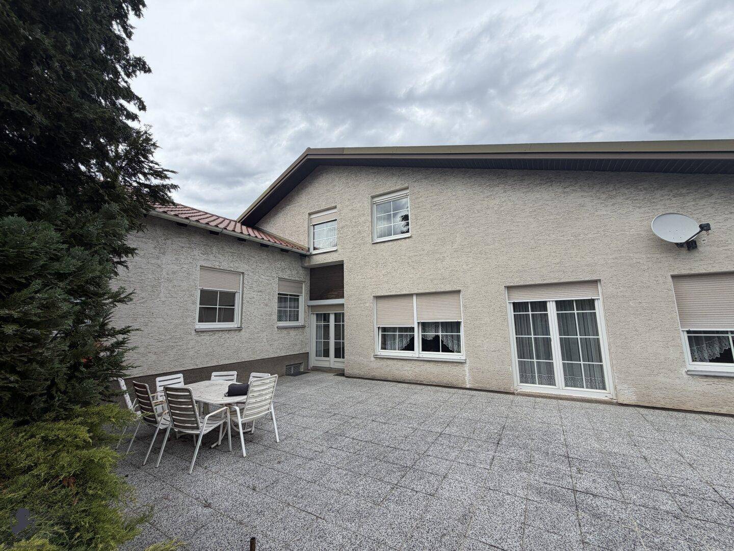 Hausfassade mit Terrasse und Gartenmöbeln, umgeben von grünen Büschen.