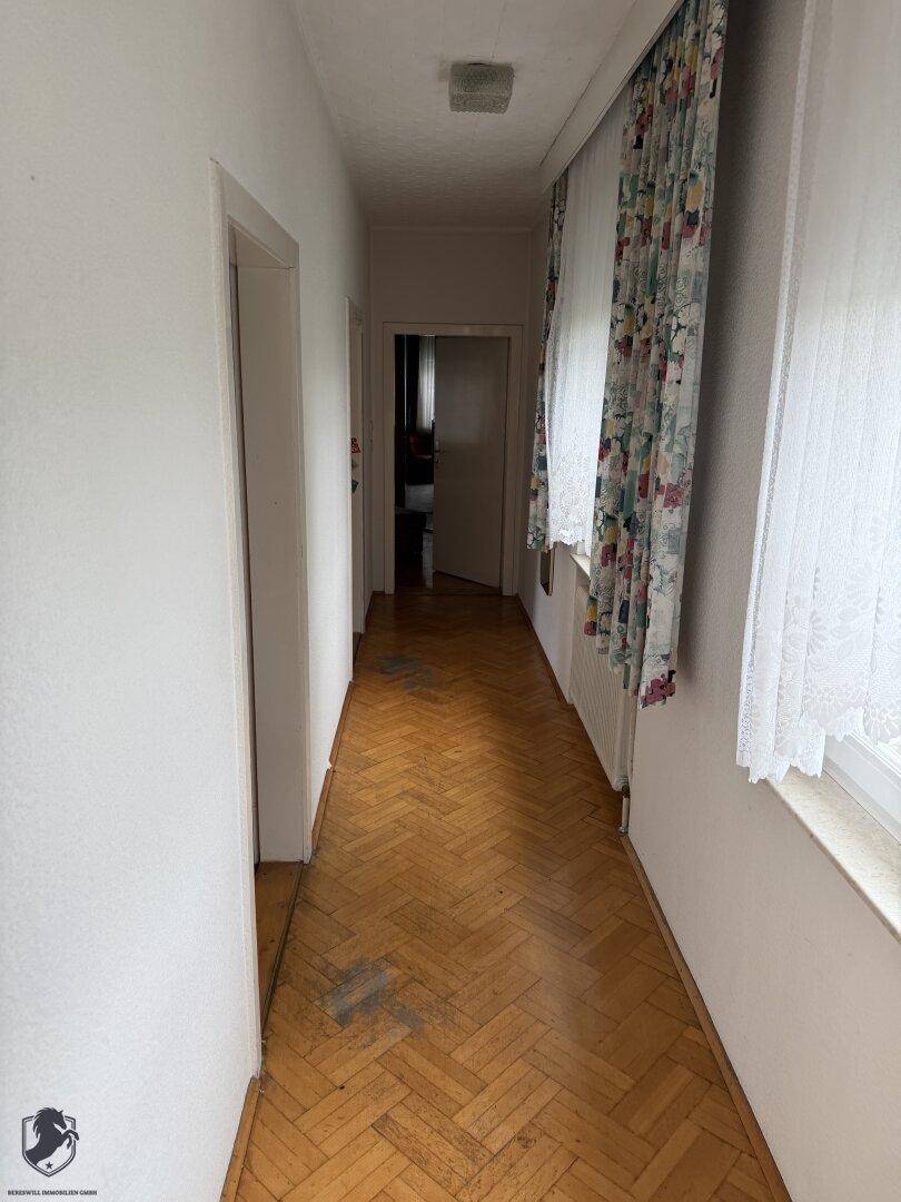 Langer Flur mit Fischgrätparkett und Fenstern, die Tageslicht hereinlassen.