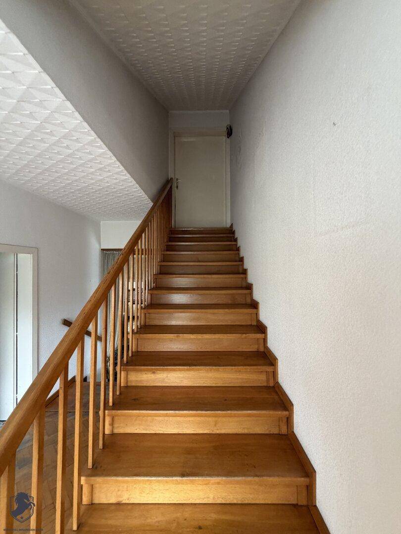 Holztreppe mit Geländer führt zu einer oberen Etage, umgeben von weißen Wänden.