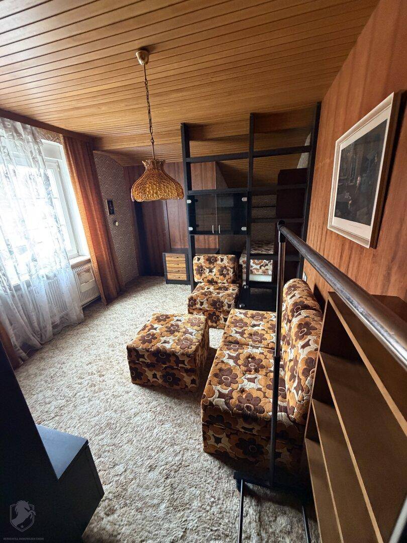 Wohnzimmer mit gemustertem Teppichboden, Holzdecke und Sitzmöbeln im 70er-Jahre-Stil.