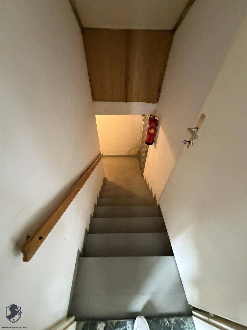 Treppenabgang in den Kellerbereich mit Betonstufen und einem Feuerlöscher an der Wand.