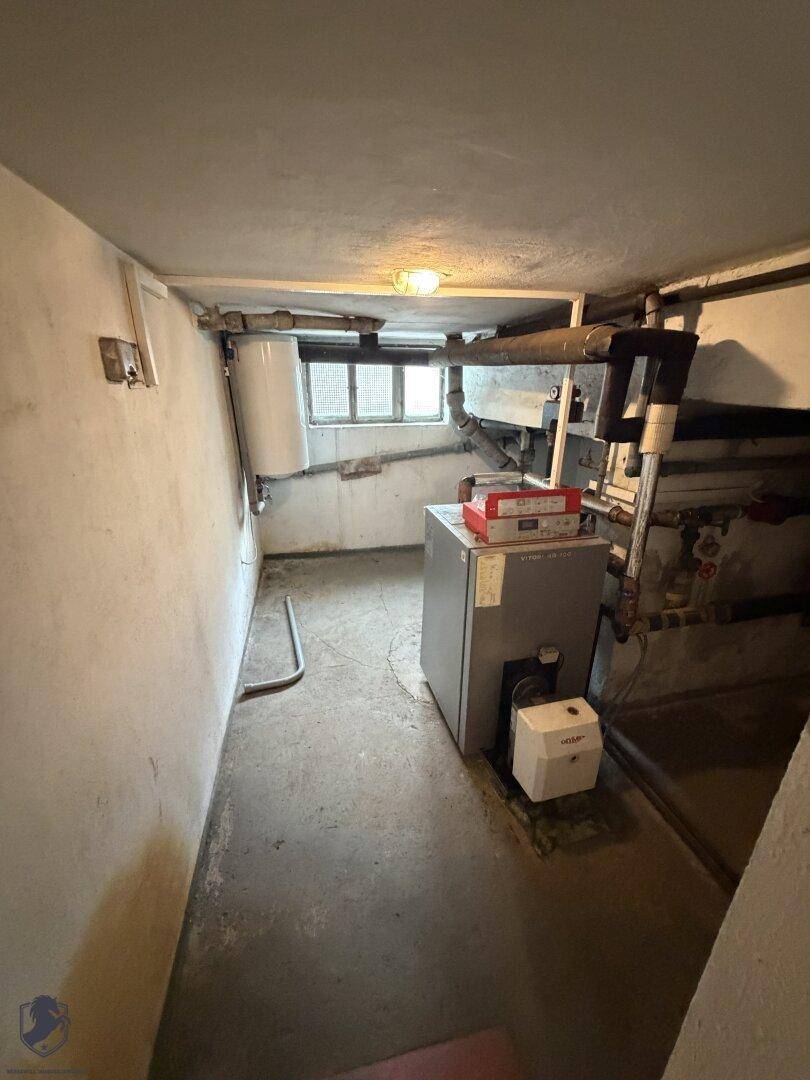 Heizungsraum im Keller mit Heizkessel, Rohren und kleinem Fenster.