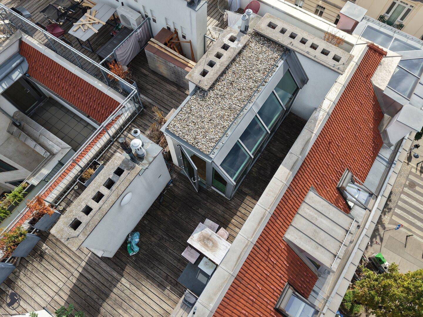 Luftaufnahme einer Dachterrasse mit Holzboden, Glasaufbau und Sitzgelegenheiten, umgeben von städtischen Dächern.