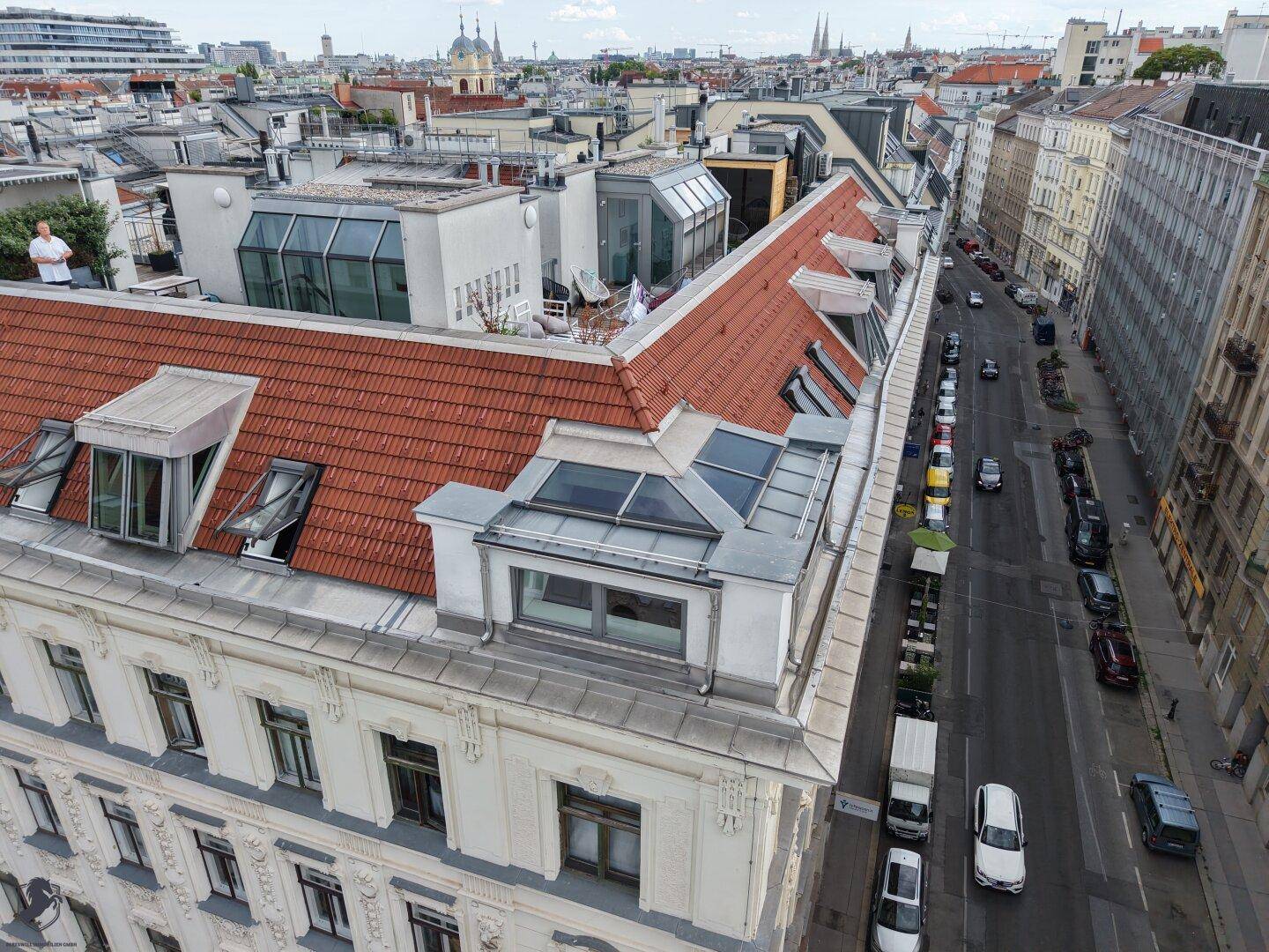 Gebäudefassade mit rotem Ziegeldach, Dachgauben und einer belebten Straße im Vordergrund, umgeben von Stadtansichten.