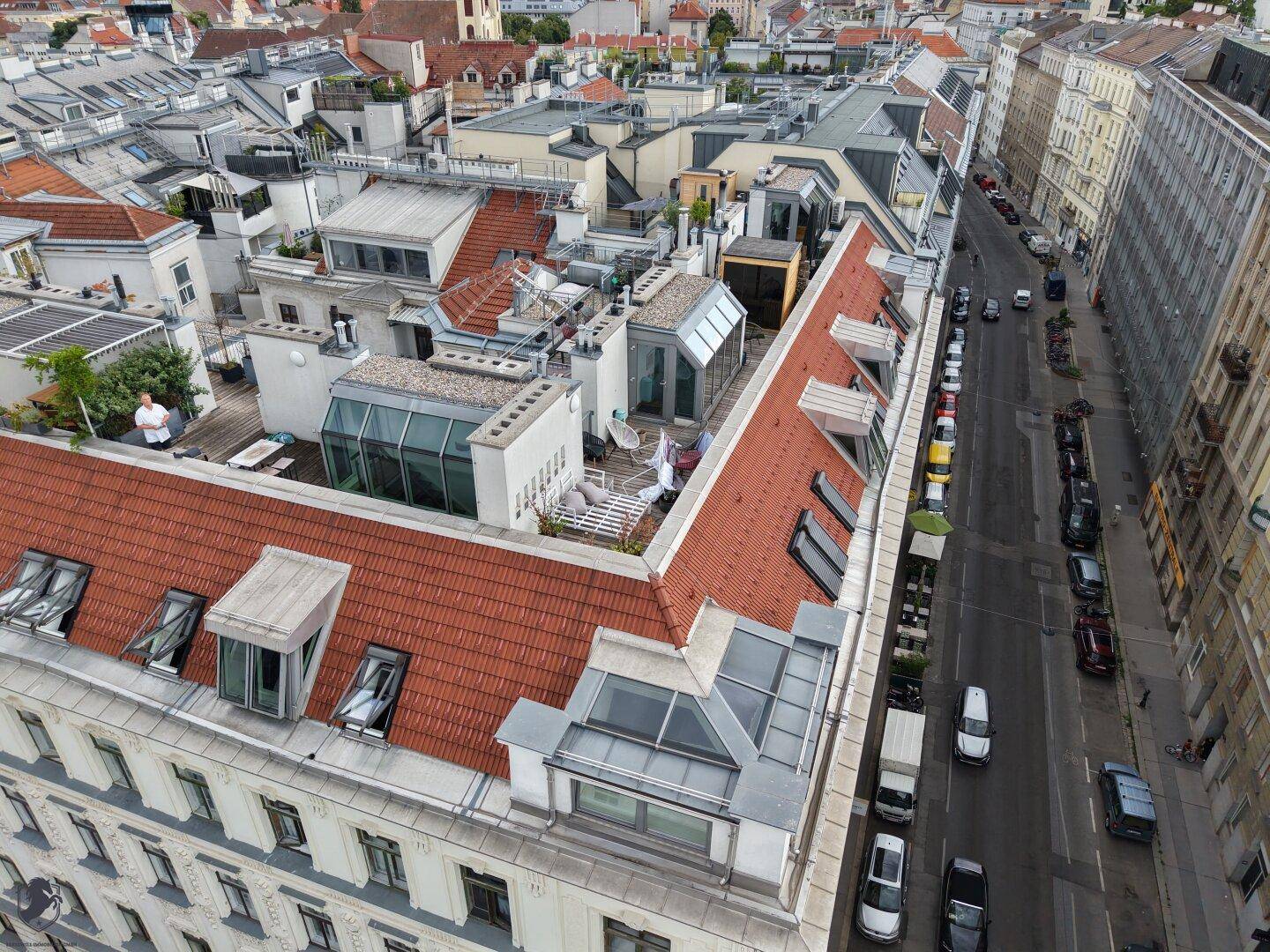Weitläufige Luftaufnahme einer Stadtstraße mit historischen Gebäuden, roten Dächern und mehreren Dachterrassen.
