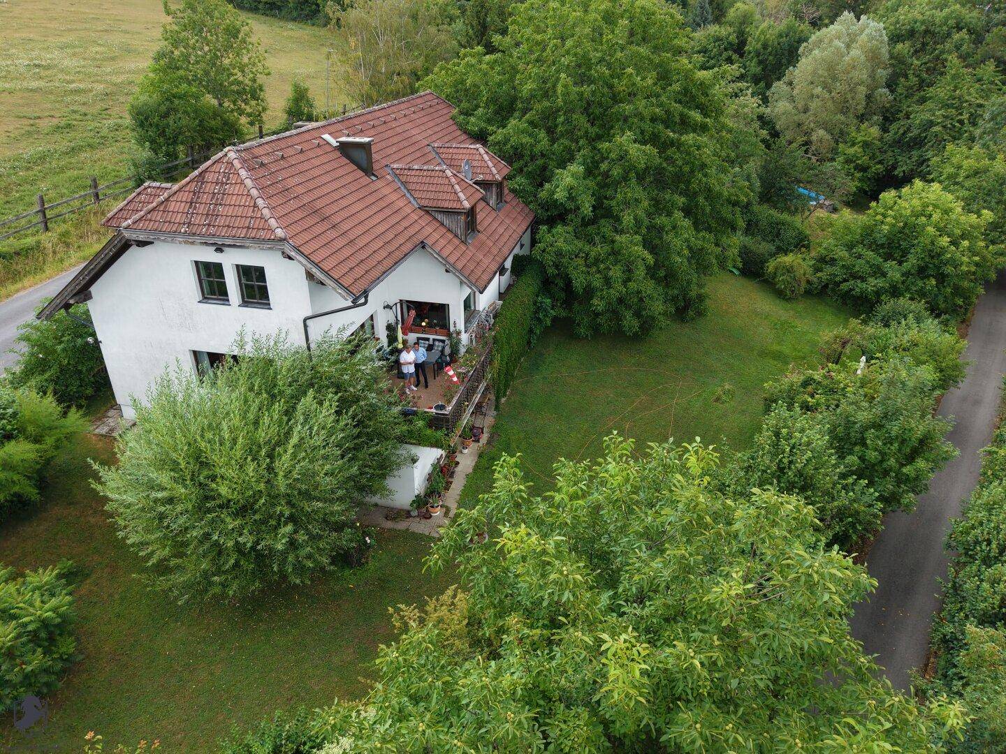 Luftaufnahme des Hauses mit bewohntem Balkon, eingebettet in eine grüne Landschaft mit Bäumen und Wiesen.