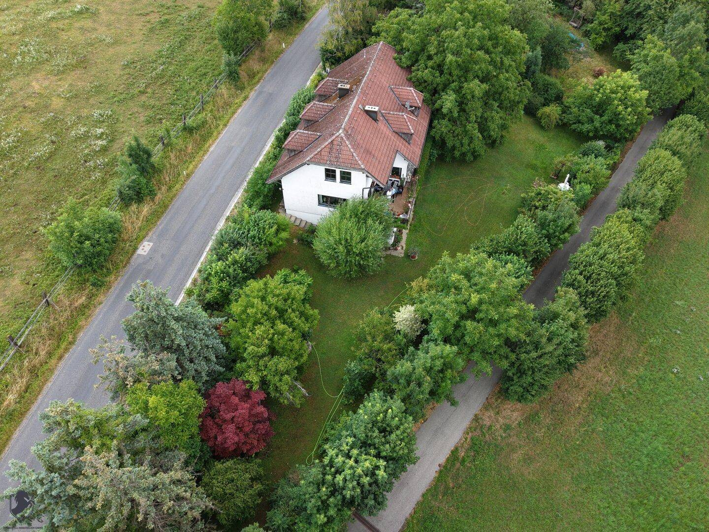 Luftaufnahme des Anwesens mit dem Haus, umgeben von viel Grün, einer Straße und weiten Feldern.