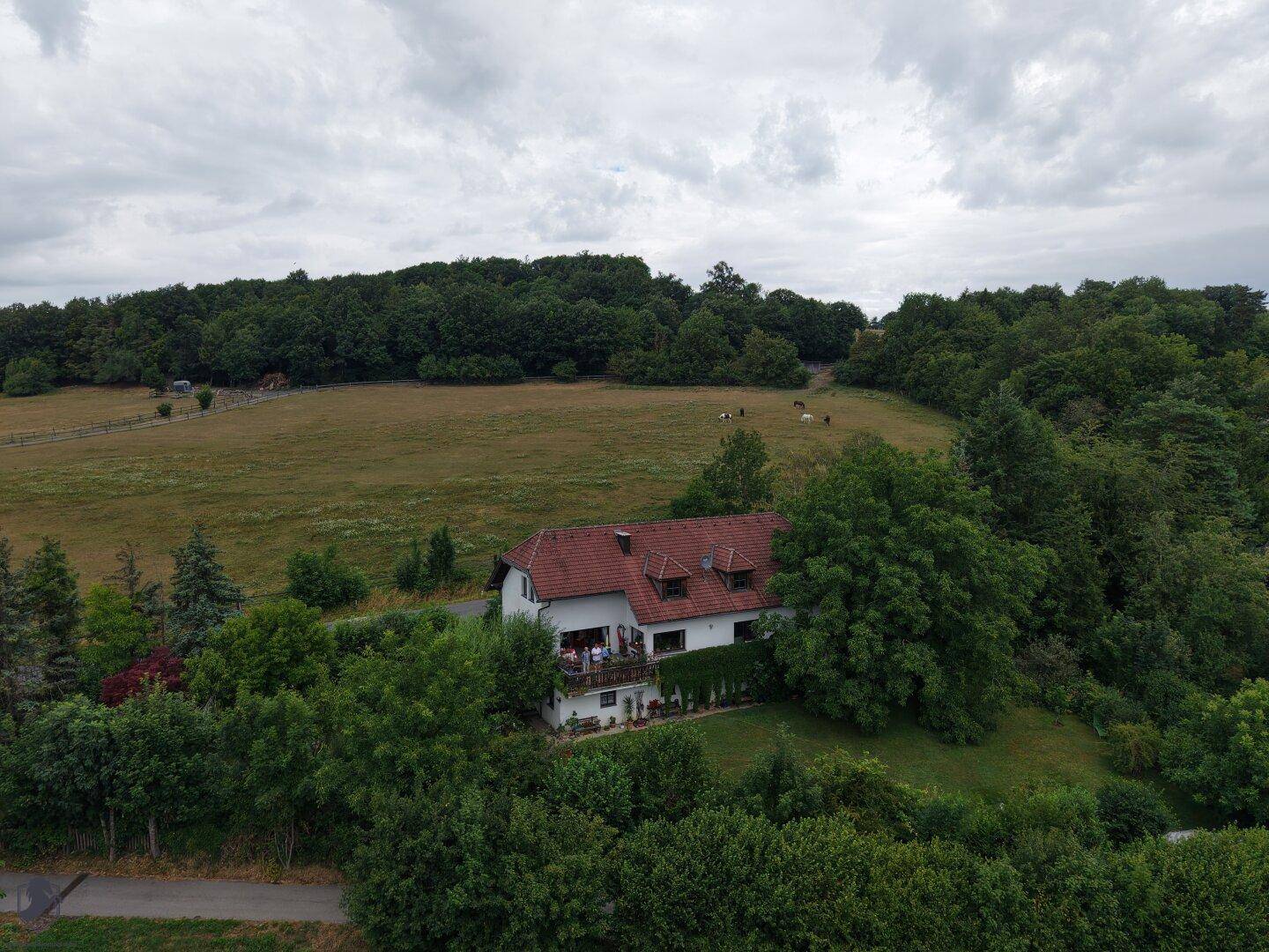 Panoramische Luftaufnahme des Hauses mit Balkon, umgeben von Wäldern und Feldern unter bewölktem Himmel.