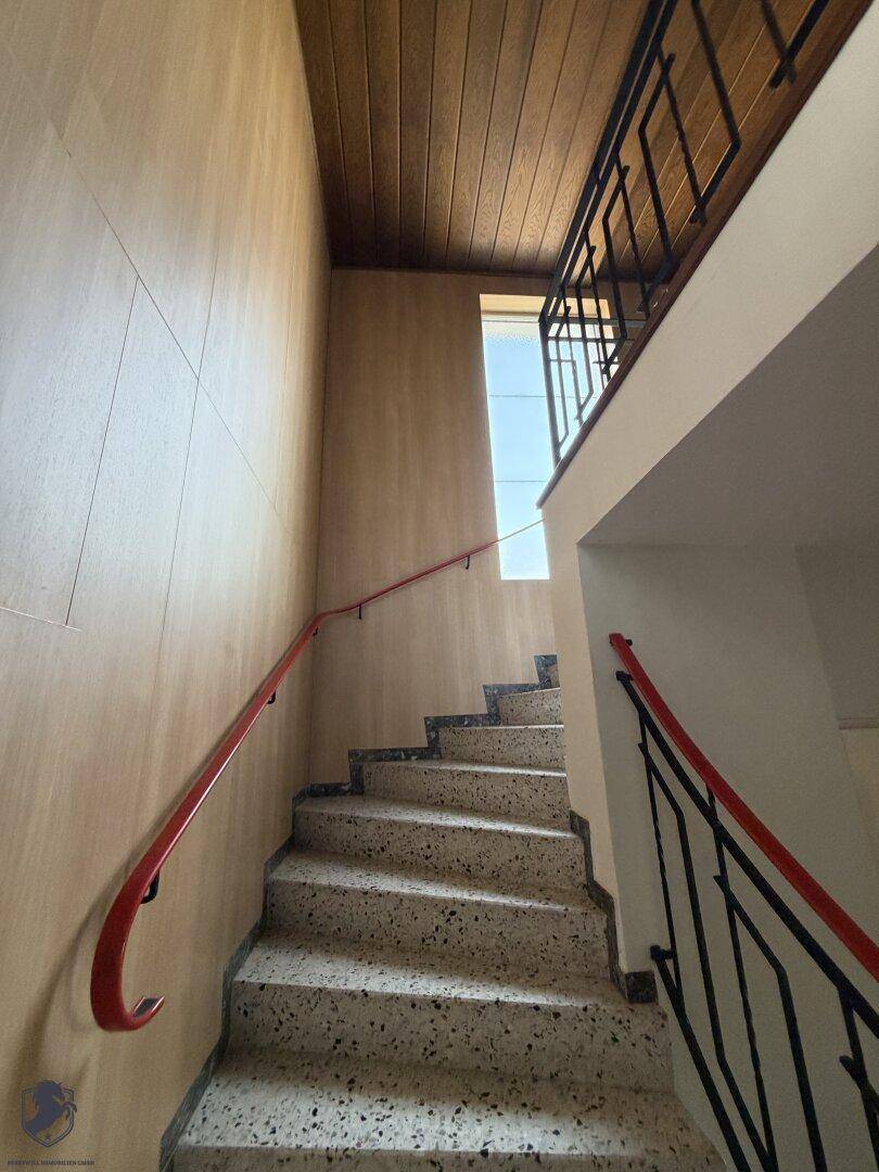 Treppenhaus mit Terrazzostufen, Holzverkleidung und rotem Handlauf.