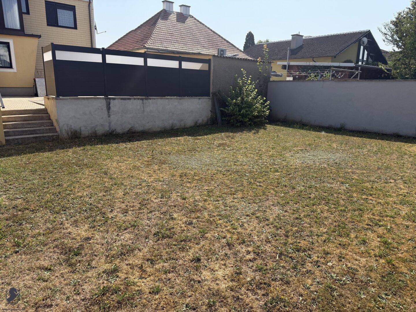 Gartenbereich mit trockenem Rasen, einer niedrigen Mauer und einem Zaun im Hintergrund.