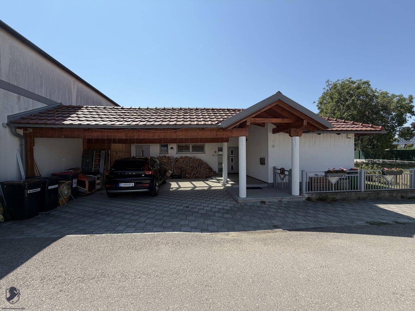 Vorderansicht des Hauses mit großem Carport und gepflasterter Einfahrt unter blauem Himmel.