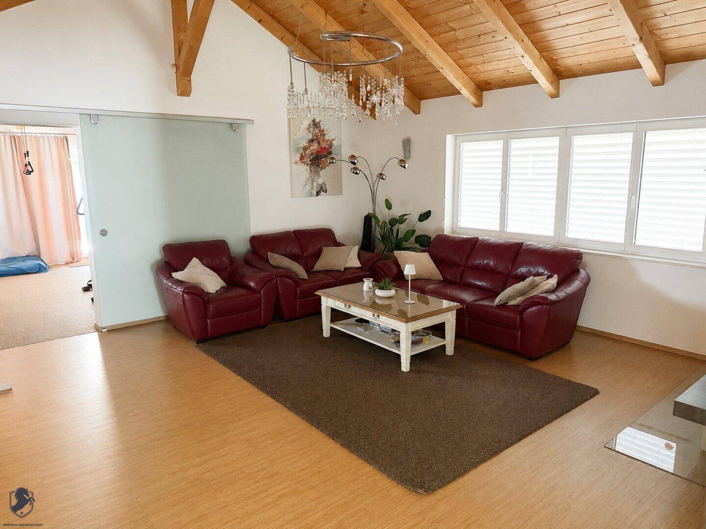 Geräumiges Wohnzimmer mit hohen Holzdecken, roten Ledersofas und großen Fenstern.