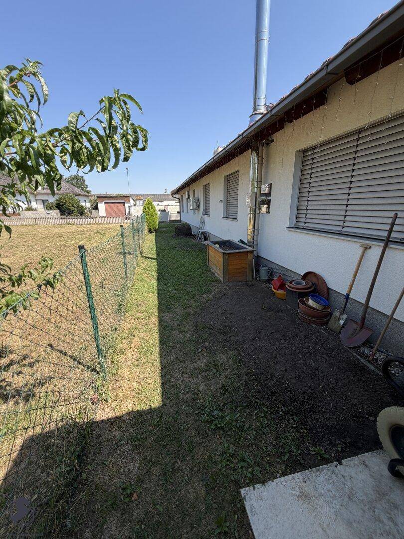 Seitenansicht des Hauses mit schmalem Rasenstreifen, Zaun und Gartengeräten.
