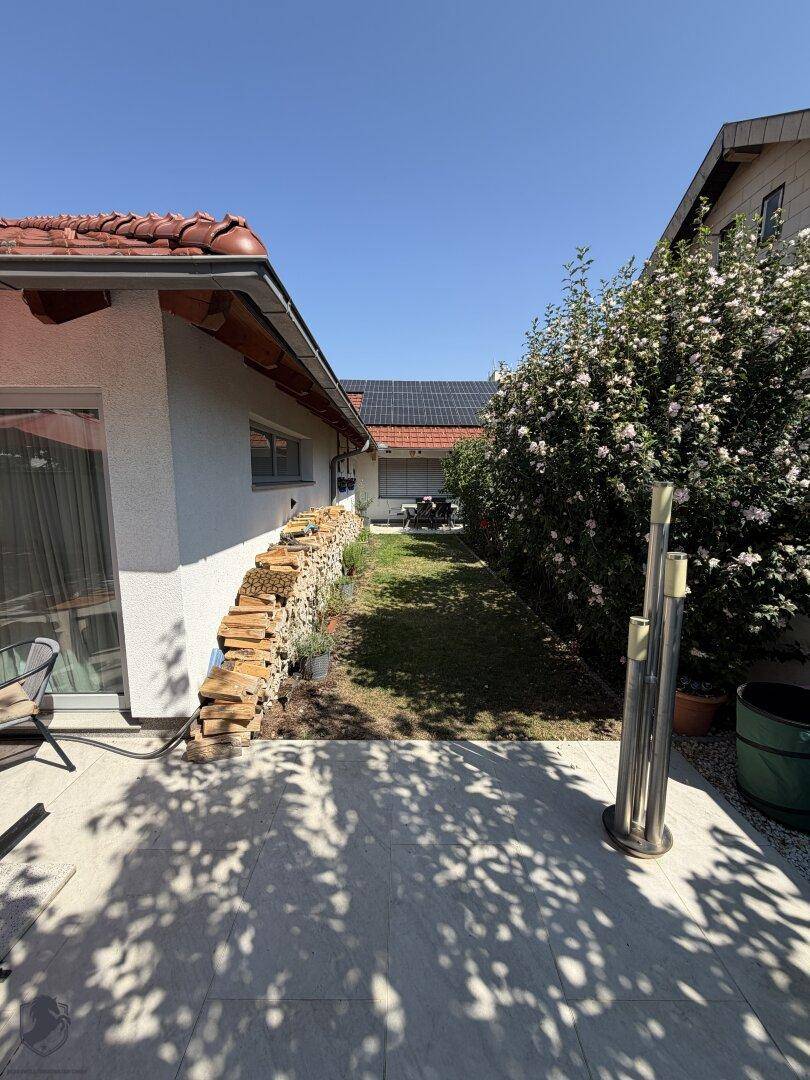Seitenansicht des Hauses mit Holzstapel, Rasenstreifen und blühendem Busch.