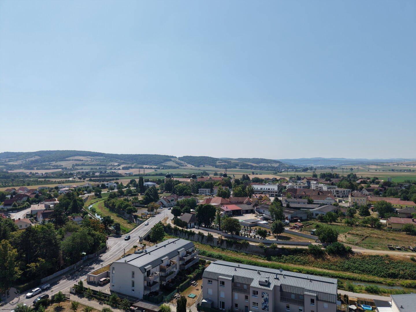 Weitläufige Luftaufnahme einer ländlichen Stadt mit grünen Hügeln und Feldern im Hintergrund.