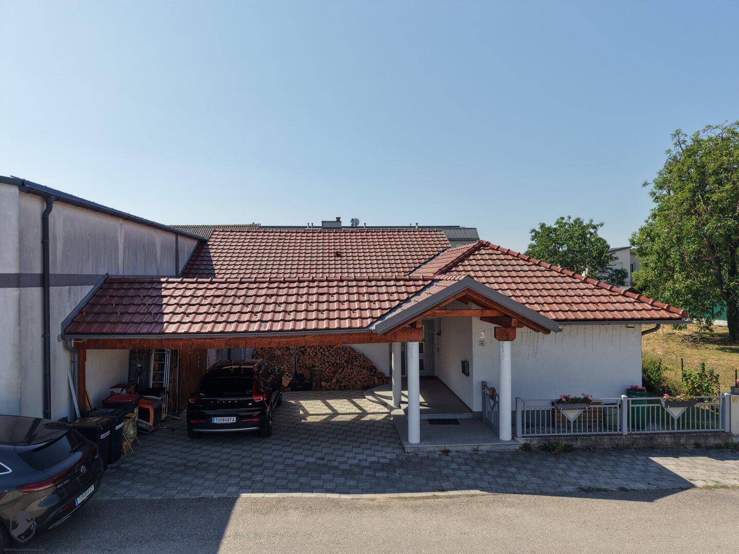 Einfamilienhaus mit überdachtem Carport, Ziegeldach und Holzstapel im Freien unter blauem Himmel.