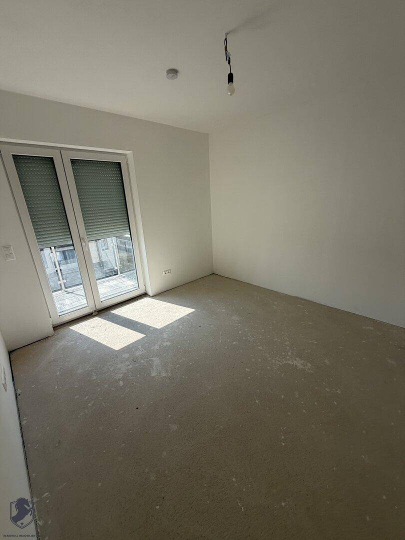 Helles, unfertiges Zimmer mit weiß gestrichenen Wänden, Rohbetonboden und Zugang zum Balkon.