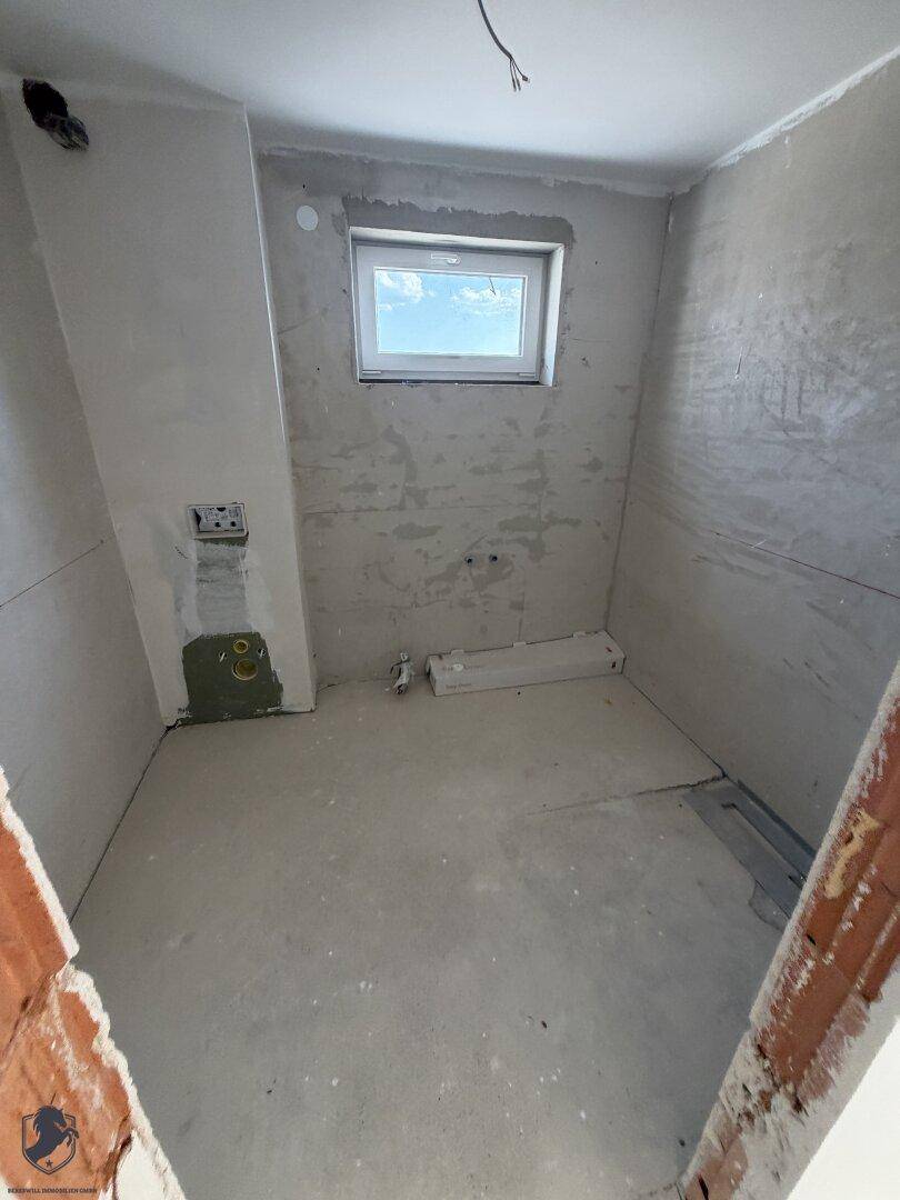 Unfertiges Badezimmer im Rohbau mit kleinem Fenster und sichtbaren Installationen für Sanitäranlagen.
