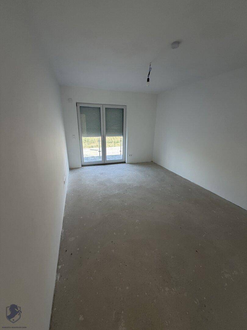 Geräumiges Zimmer im Rohbau mit weiß gestrichenen Wänden und großem Fenster mit Rollläden.