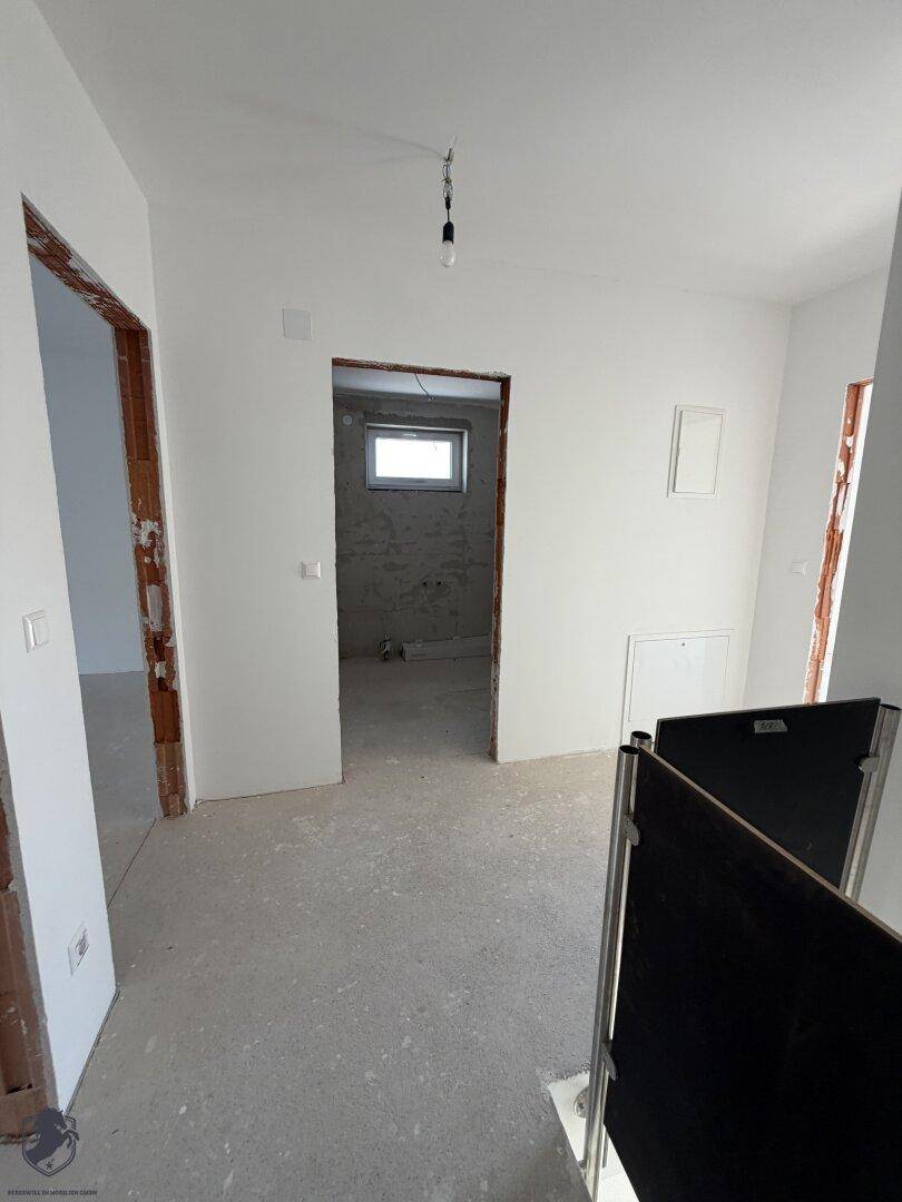 Flur im Rohbau mit weiß gestrichenen Wänden, Betonboden und Zugang zu weiteren Räumen.