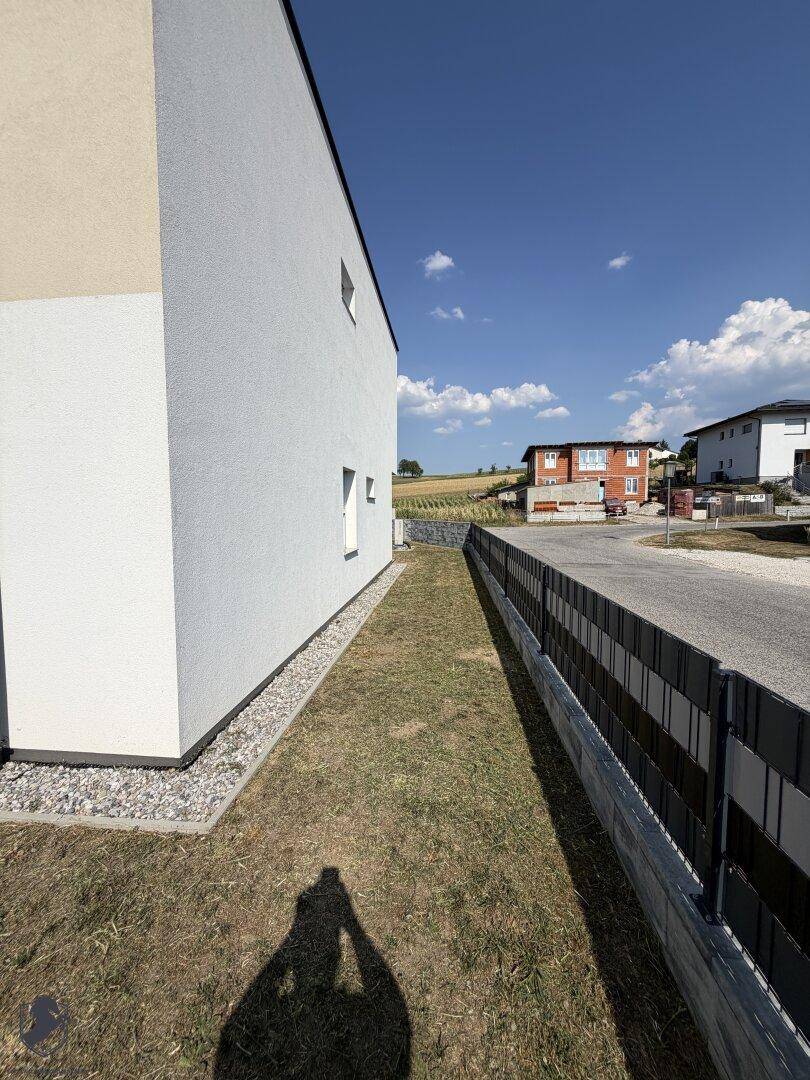 Seitenansicht des Neubaus mit schmalem Rasenstreifen, Zaun und Blick auf die Landschaft.