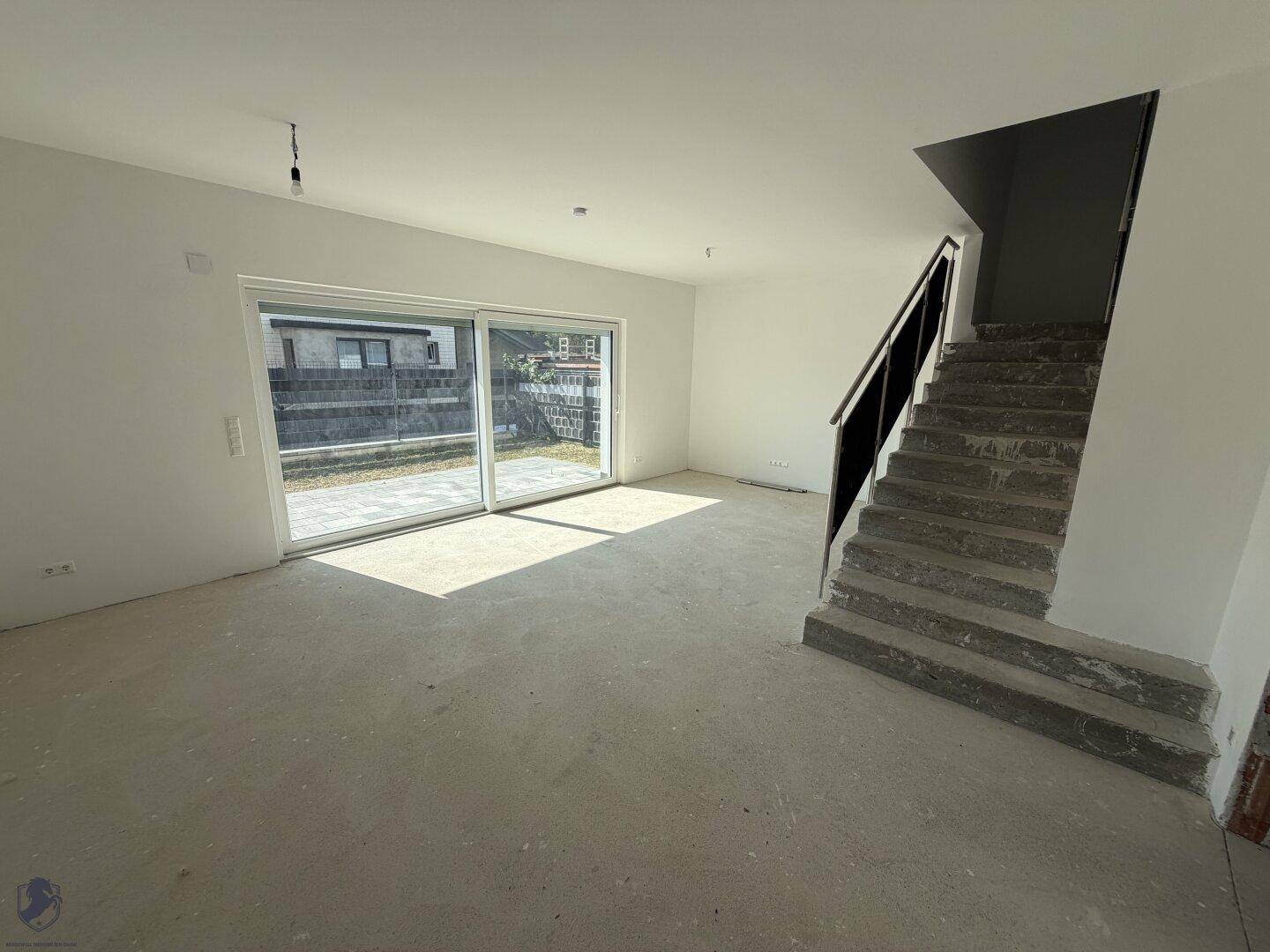 Geräumiges Wohnzimmer im Rohbau mit Betonboden, weiß gestrichenen Wänden und Gartenzugang.