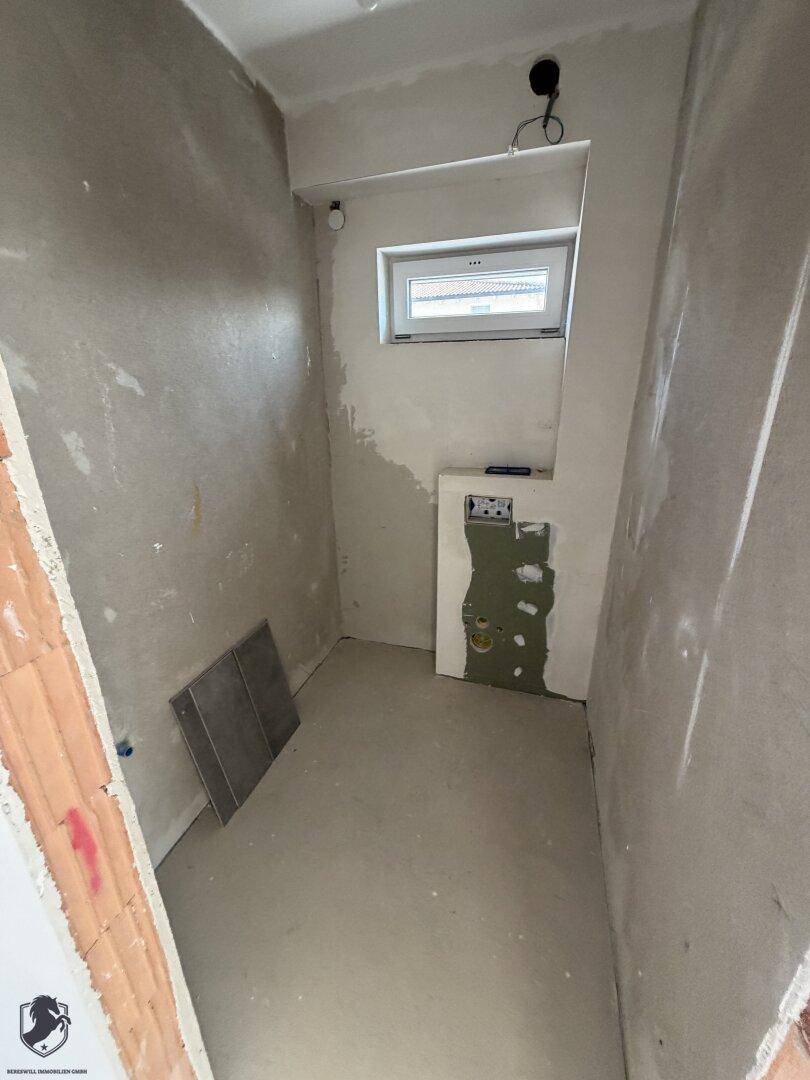 Unfertiges Badezimmer im Rohbau mit kleinem Fenster und vorbereiteten Anschlüssen für Sanitär.