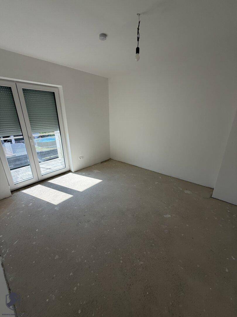 Helles Zimmer im Rohbau mit weiß gestrichenen Wänden, Betonboden und Zugang zum Balkon.