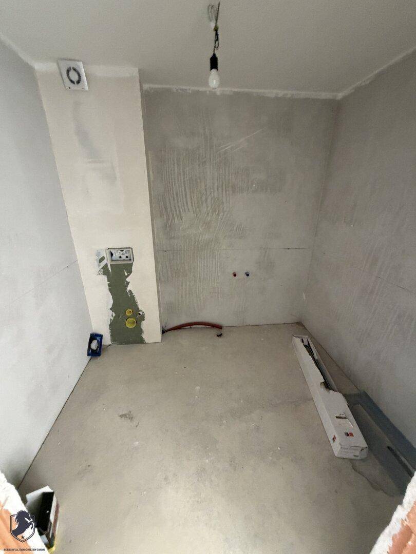 Unfertiges Badezimmer im Rohbau mit sichtbaren Installationen und einem Abluftventilator.
