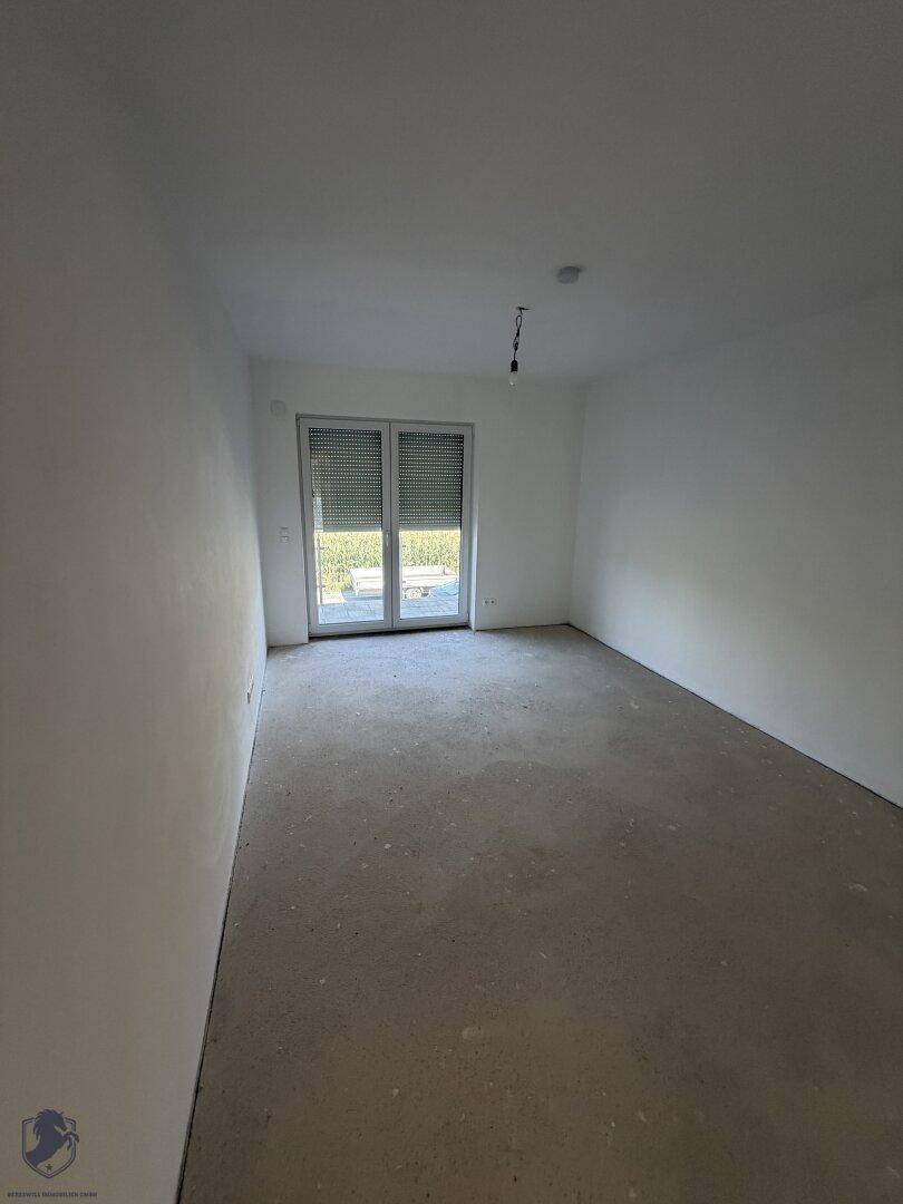 Geräumiges Zimmer im Rohbau mit weiß gestrichenen Wänden und großem Fenster mit Rollläden.