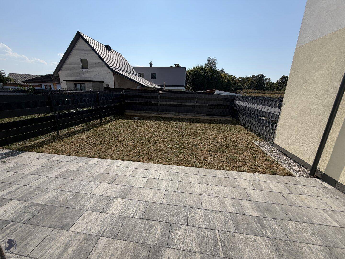 Gepflasterte Terrasse mit angrenzendem Gartenbereich und Blick auf Nachbarhäuser.