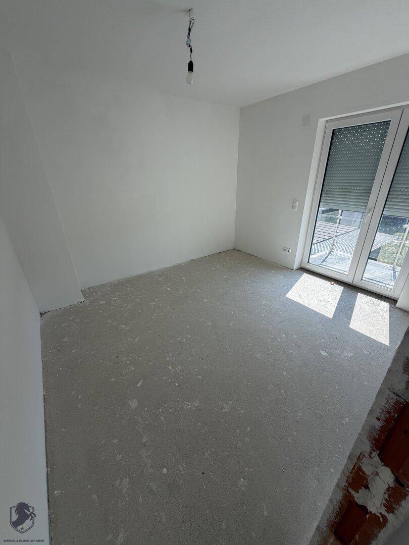 Unfertiges Schlafzimmer mit Rohbetonboden, weißen Wänden und Zugang zu einem Balkon.