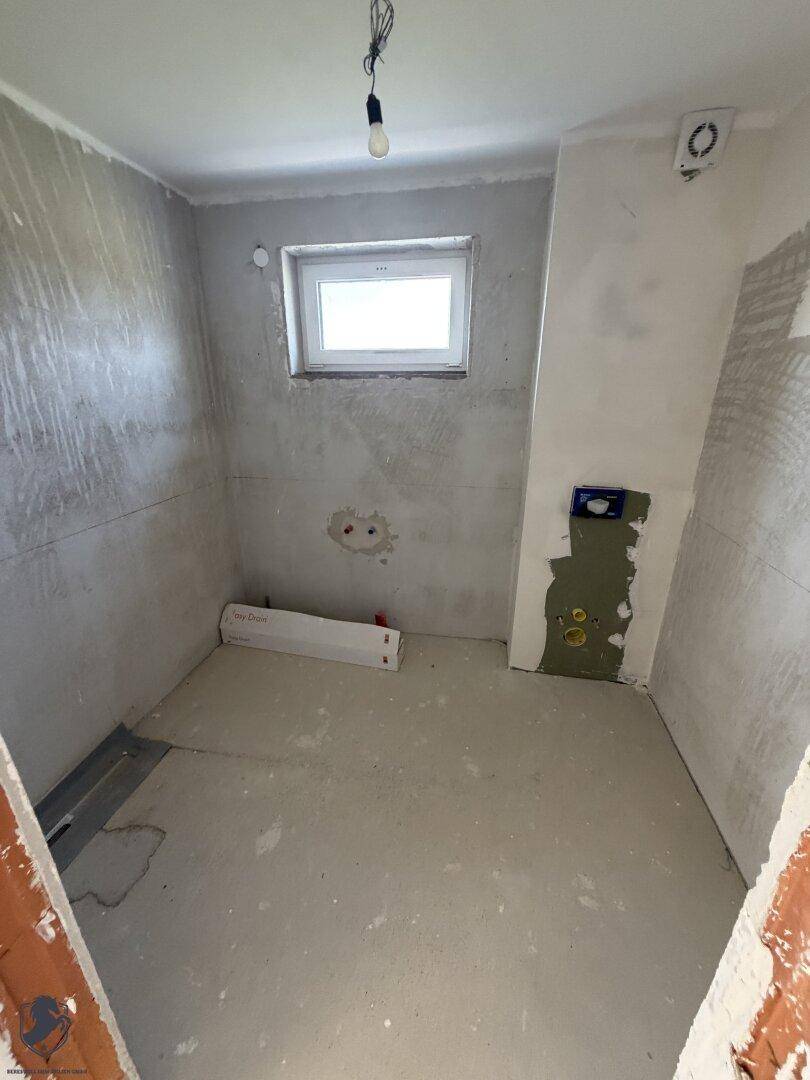 Unfertiges Badezimmer mit kleinem Fenster und vorbereiteten Anschlüssen für Sanitäranlagen.