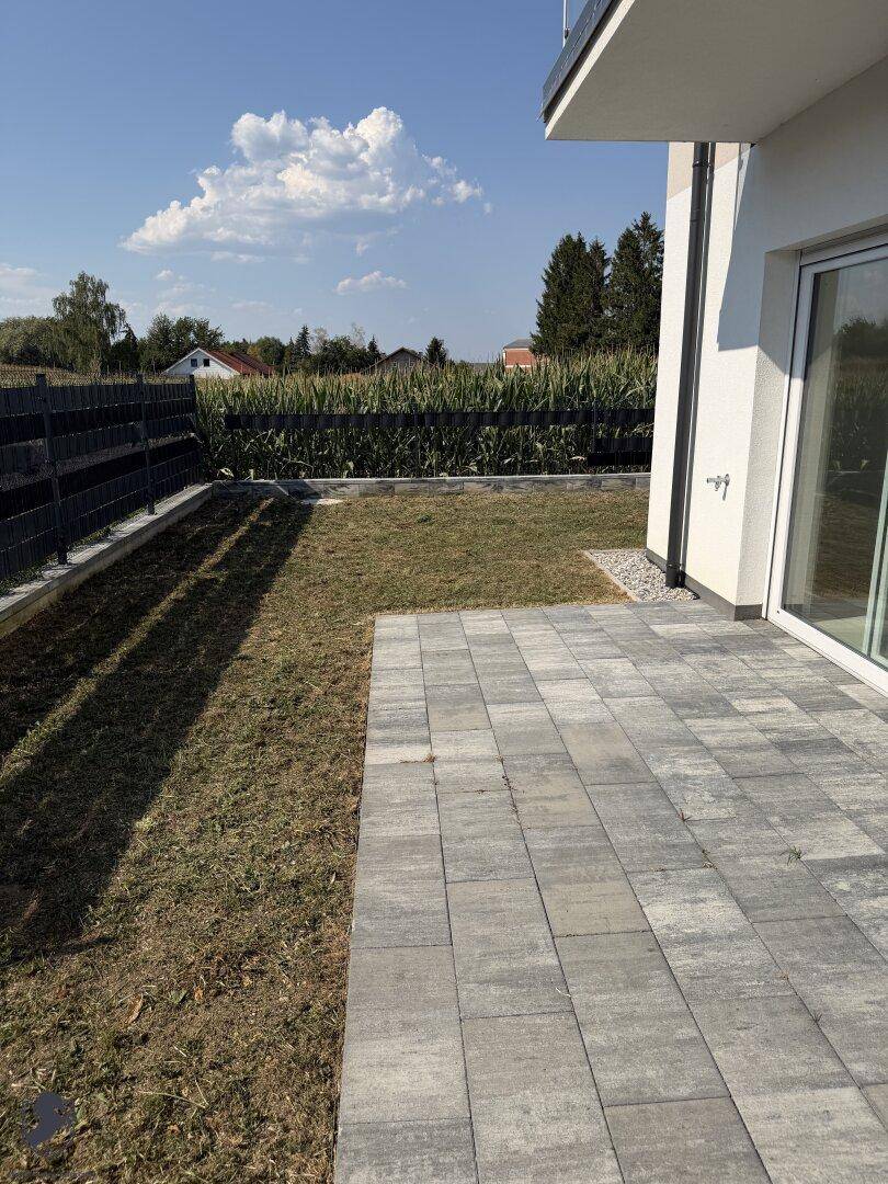 Gepflasterter Terrassenbereich mit Rasenfläche und Blick auf angrenzende Felder.