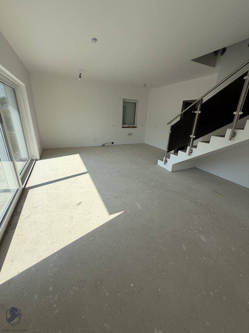 Geräumiger, unfertiger Wohnbereich mit Rohbetonboden und viel Tageslicht durch große Fenster.