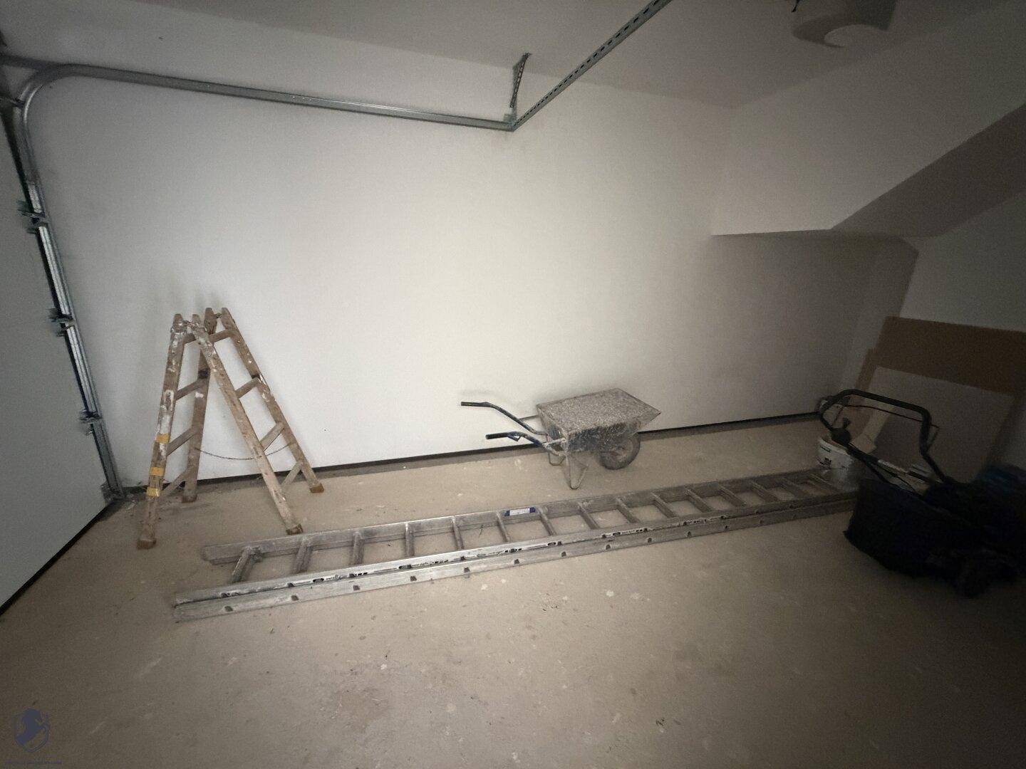 Unfertige Garage mit Rolltor, Betonboden und gelagerten Baugeräten.