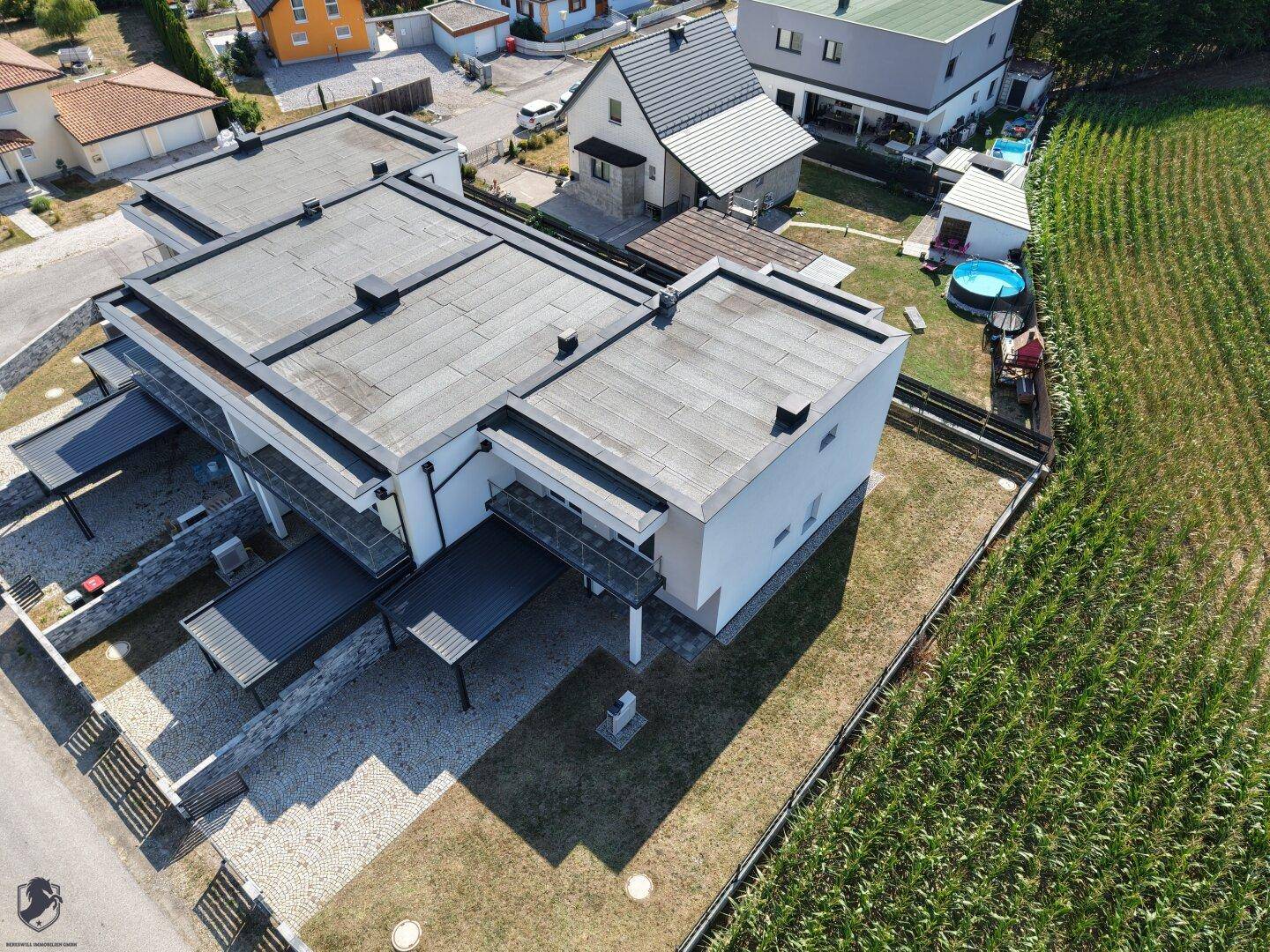Moderne Reihenhäuser mit Flachdächern und Carports, angrenzend an ein Maisfeld unter blauem Himmel.