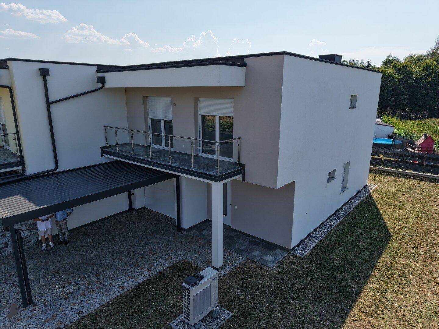 Seitenansicht eines modernen Reihenhauses mit Balkon, Carport und sichtbarer Wärmepumpe im Gartenbereich.