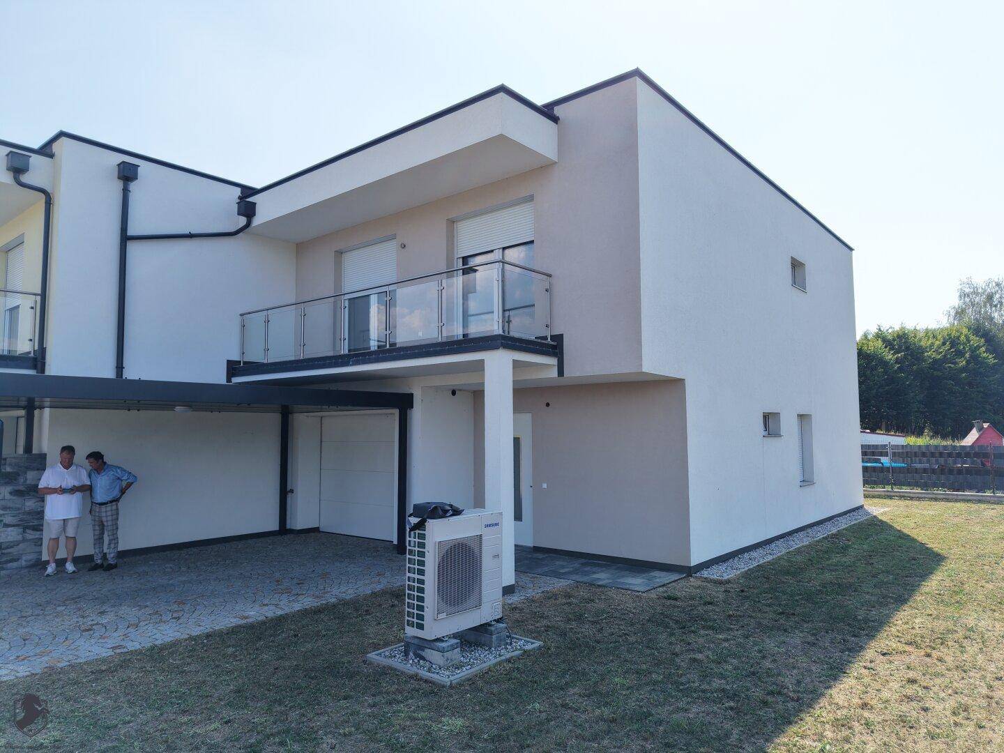 Modernes Reihenhaus mit Balkon, Carport und Wärmepumpe in sonniger Umgebung mit Rasenfläche.