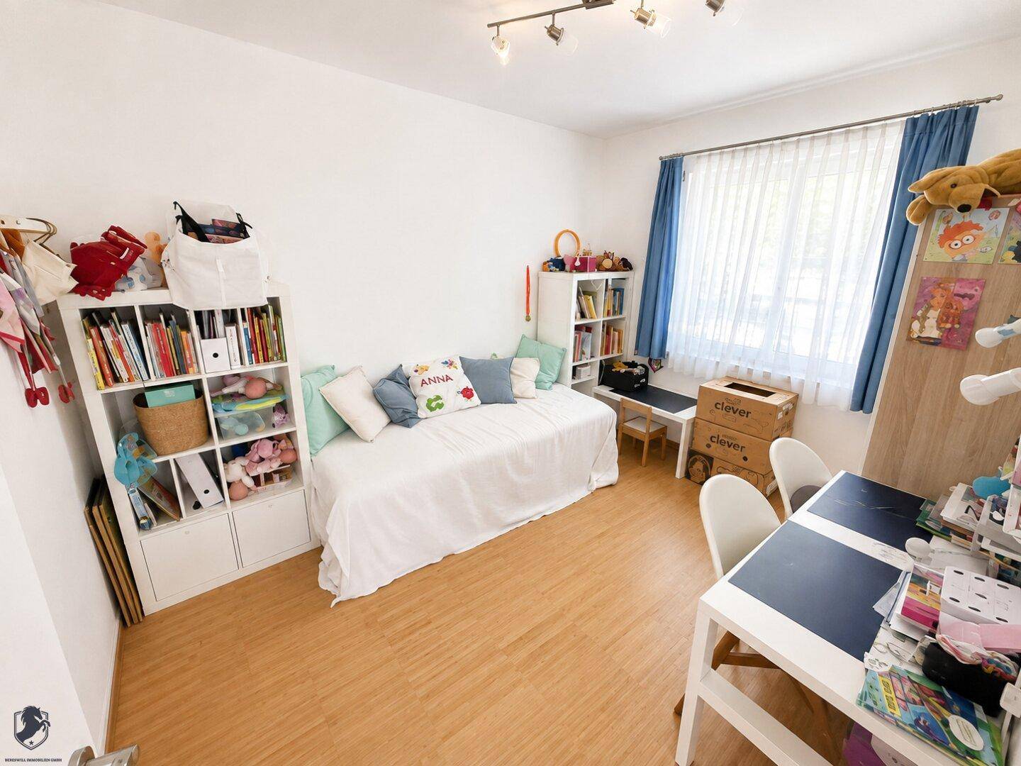 Kinderzimmer mit Tagesbett, Bücherregalen und Schreibtisch, hell beleuchtet durch ein Fenster.