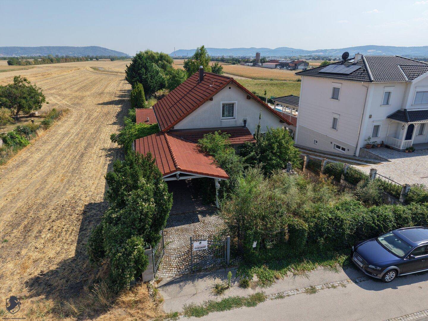 Luftaufnahme der Vorderseite des Hauses mit Carport, gepflasterter Einfahrt und angrenzendem Feld.