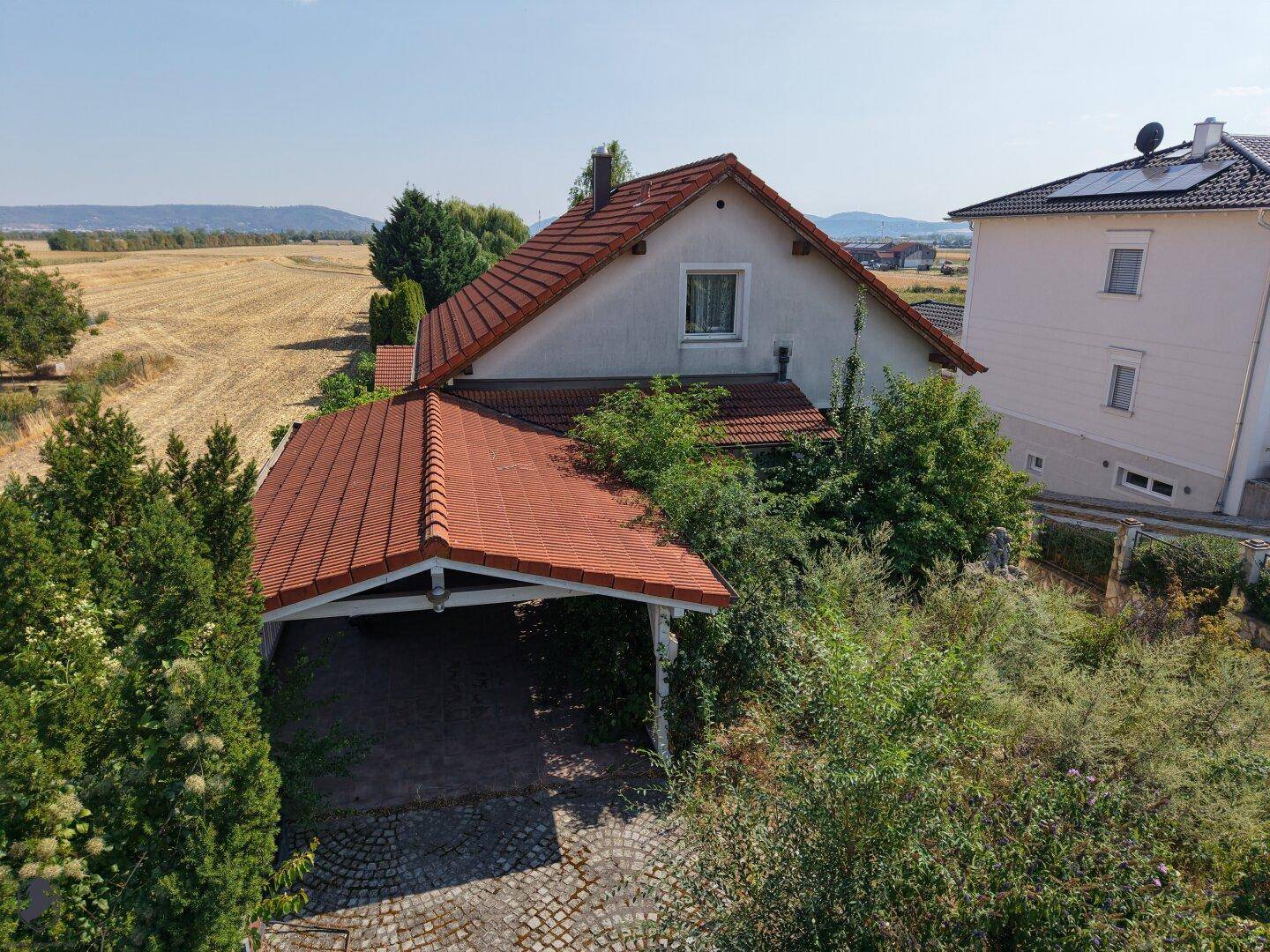 Luftaufnahme der Hausvorderseite mit Carport und dem weiten, angrenzenden Feld unter blauem Himmel.