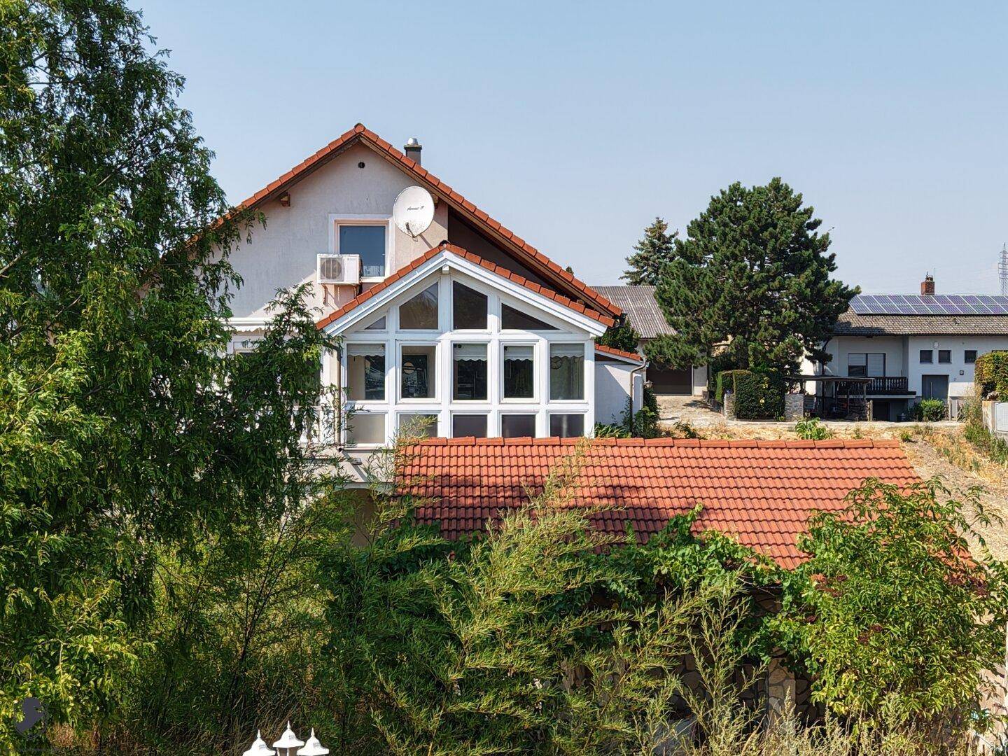 Ansicht des Hauses mit verglastem Wintergarten und Balkon, umgeben von üppigem Grün.