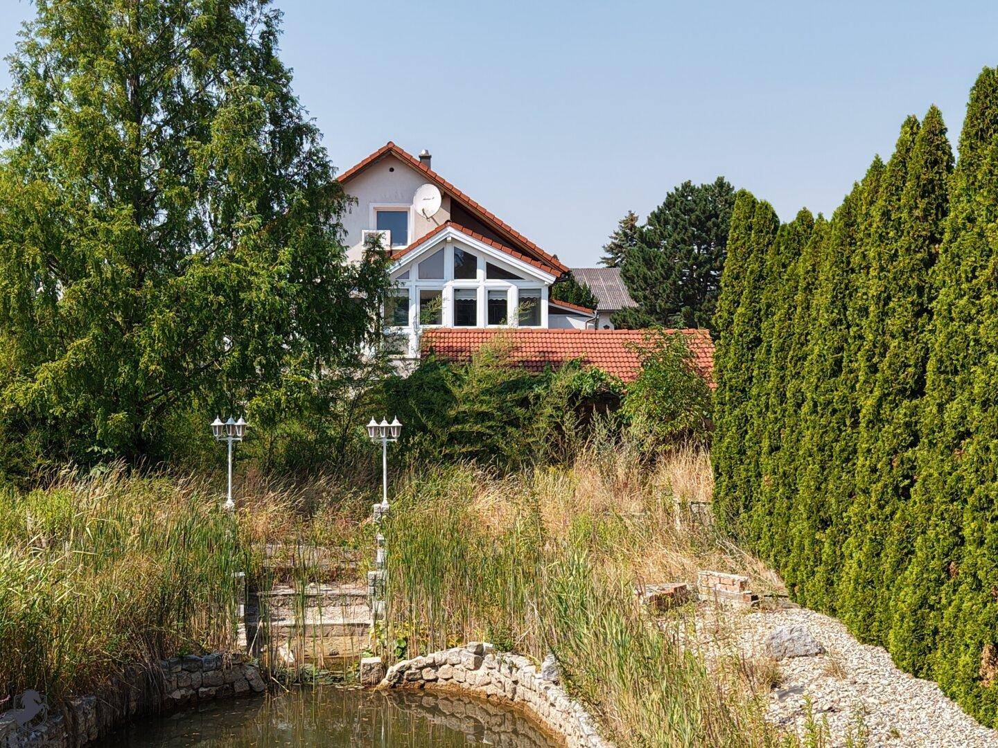 Blick auf den Teich im Vordergrund, mit dem Haus und der Hecke im Hintergrund unter blauem Himmel.