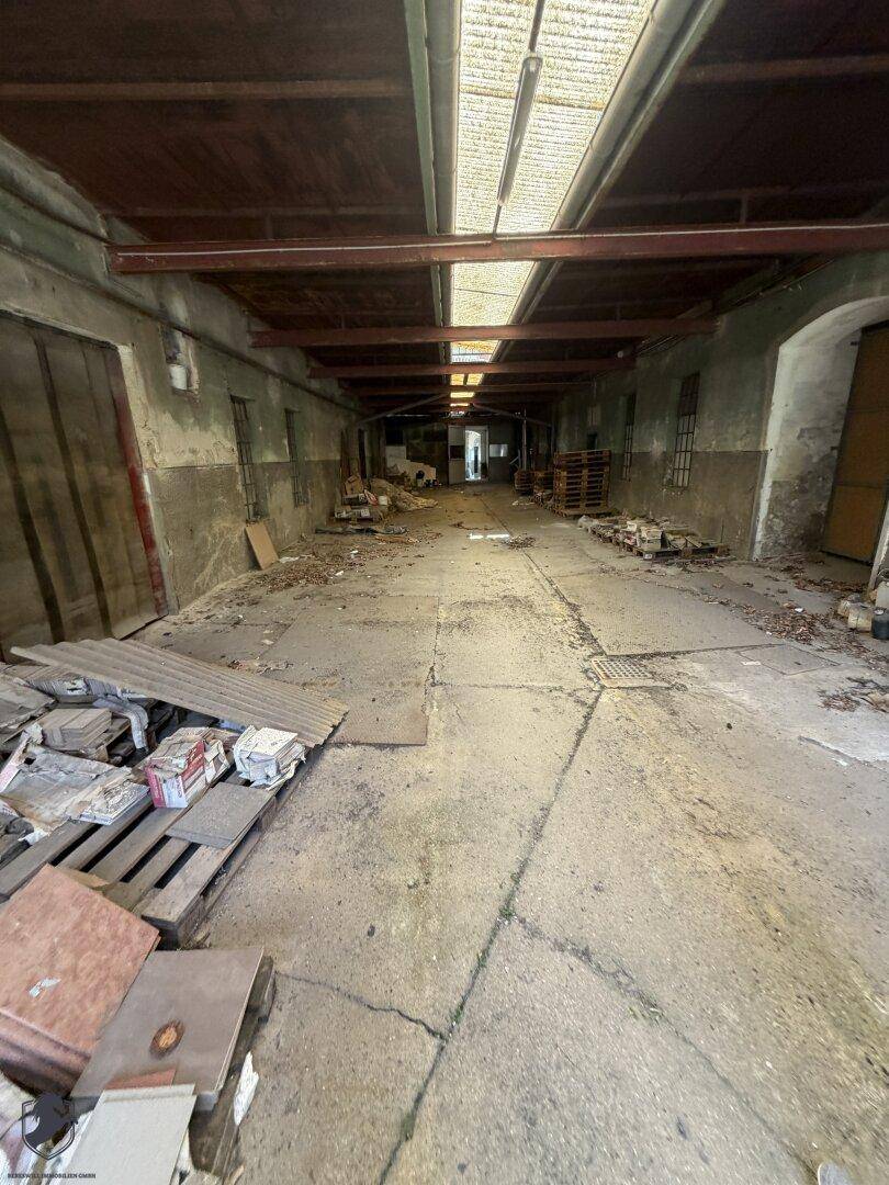 Langer, dunkler und verwahrloster Innenraum, möglicherweise eine alte Lagerhalle mit Schutt und Paletten.
