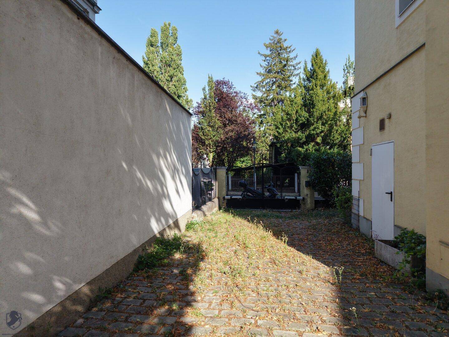 Gepflasterter Hinterhofbereich zwischen zwei Gebäuden mit Bäumen und Sträuchern im Hintergrund.