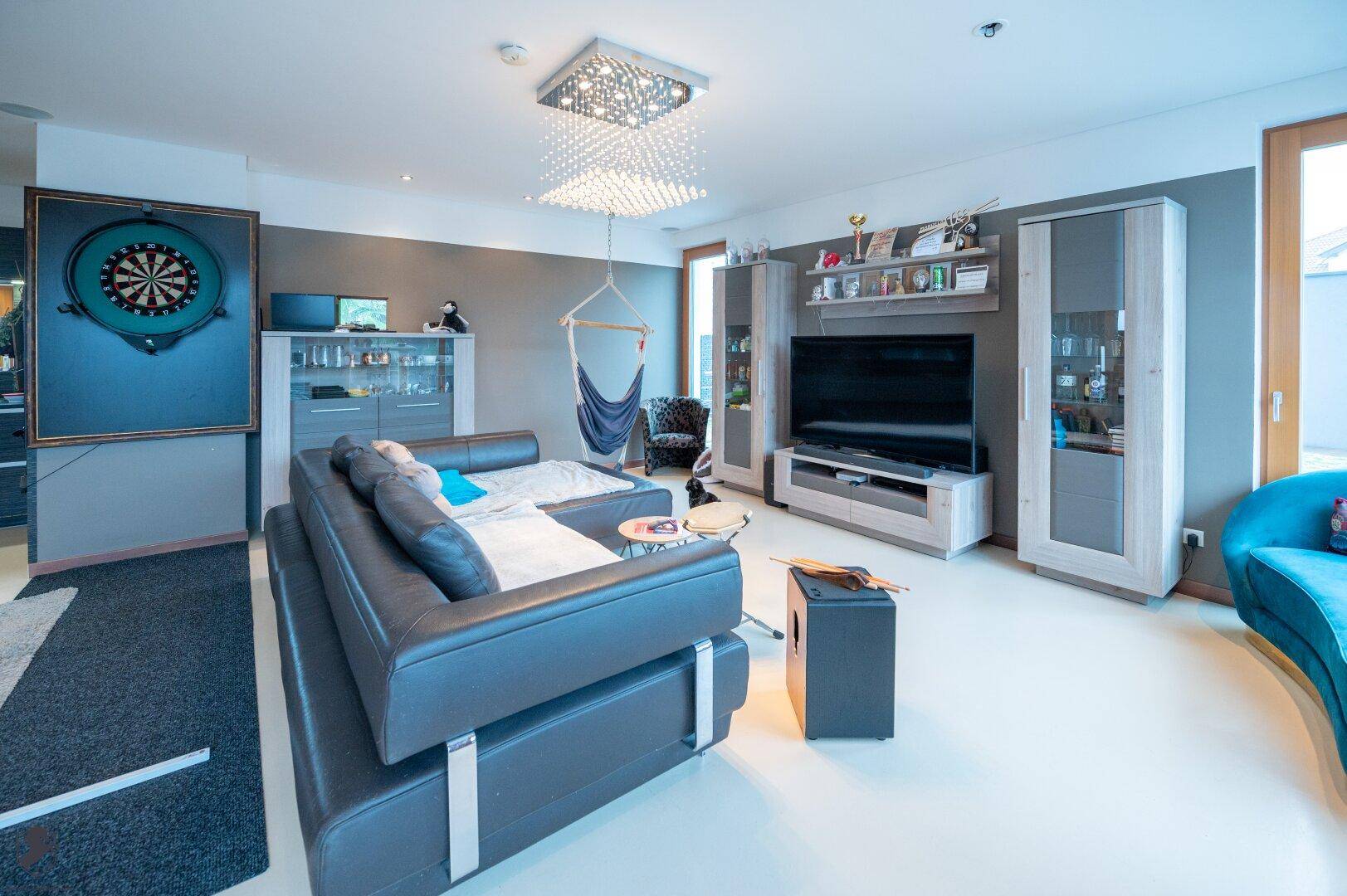 Modernes Wohnzimmer mit bequemer Sofalandschaft, großem Fernseher und einem Dartboard als Freizeitelement.